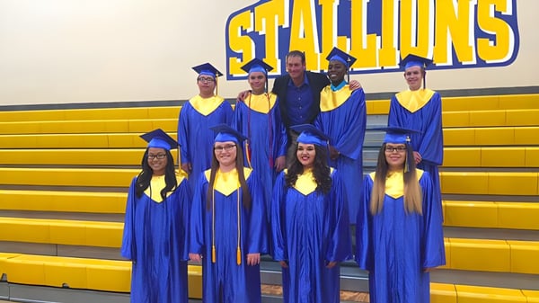 Un grupo de graduados en togas azules se encuentra frente al cartel de Stallions en las gradas amarillas de la Sequoia Charter High School.