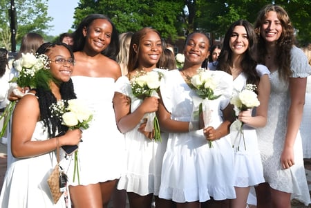 Un grupo de alumnas de la Shady Side Academy en vestidos blancos con ramos de flores está en un jardín con árboles y áreas verdes.