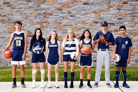Un grupo de jóvenes atletas está con equipos deportivos frente a una pared de piedra en el terreno de la Shalom Christian Academy.