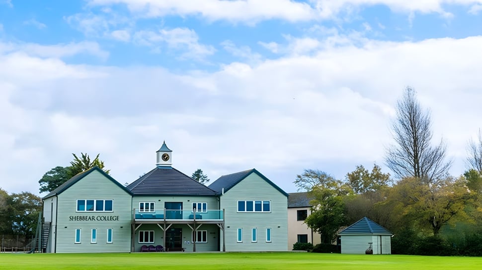 El colorido edificio de dos pisos con reloj del Shebbear College se encuentra en un campo de césped rodeado de árboles otoñales.