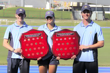 Tres personas con ropa uniforme y escudos rojos están en el campo deportivo del Sheldon College.