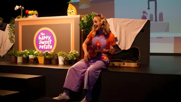 Una mujer toca la guitarra en el escenario del Sheldon College frente a una decoración de Happy Couch Potato.