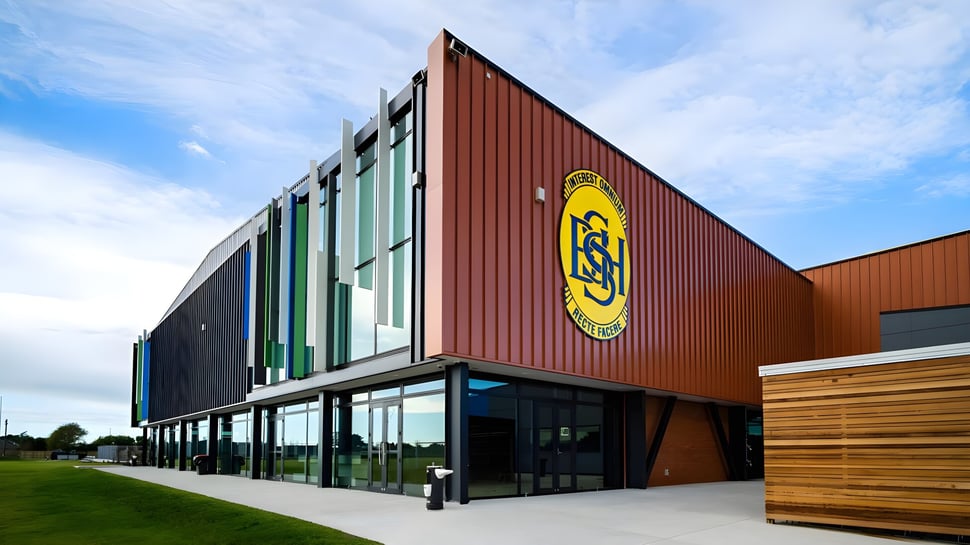 El edificio multicolor con el gran logo amarillo de Shirley Boys' High School se encuentra en un terreno cubierto de hierba bajo un cielo azul.