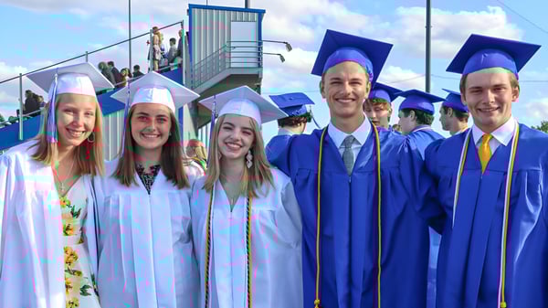 Un grupo de graduadas y graduados de la Sir Guy Carleton Secondary School en togas azules está frente a un edificio contenedor y un cielo nublado.