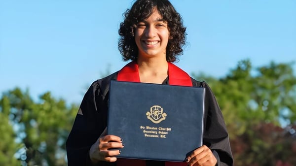 Un estudiante de la Sir Winston Churchill Secondary School lleva una toga de graduación negra y sostiene un diploma frente a un fondo con árboles y cielo azul.