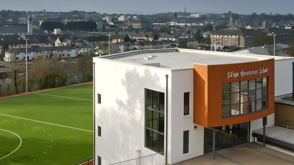 El moderno edificio de la Sligo Grammar School con grandes ventanas da a un verde campo de fútbol frente a un paisaje urbano.