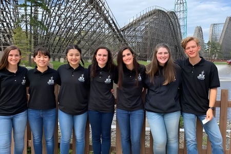 Un grupo de siete estudiantes de la Smithtown Christian School está frente a una gran montaña rusa en un parque de atracciones.