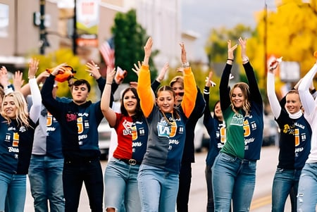 Un grupo de jóvenes en Snow College está frente a colorido follaje otoñal con los brazos levantados en pose de celebración.