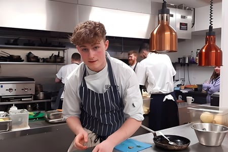 Un estudiante en uniforme de cocina está en la cocina del campus del South Downs College rodeado de utensilios de cocina.