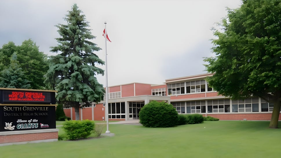 El edificio de ladrillo de la South Grenville District High School con una bandera canadiense y rodeado de árboles y césped.