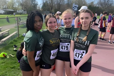 Grupo de estudiantes de la South Grenville District High School en uniformes verdes y amarillos que están en el aire libre sobre un camino pavimentado.
