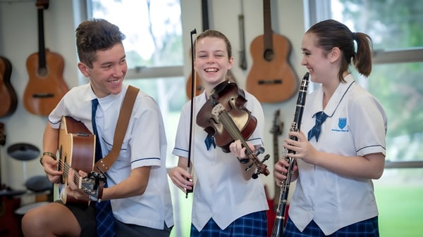 Tres estudiantes de la Southport State High School tocan guitarras en una sala con guitarras colgadas en las paredes.
