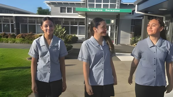 Tres estudiantes en uniformes grises están de pie frente a un edificio con un letrero verde en el campus del Spotswood College.
