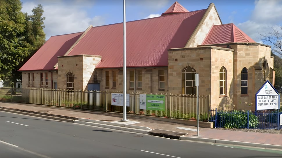 El gran edificio con techo rojo y arcos de piedra en el campus del Springbank Secondary College está rodeado por una cerca y áreas verdes.