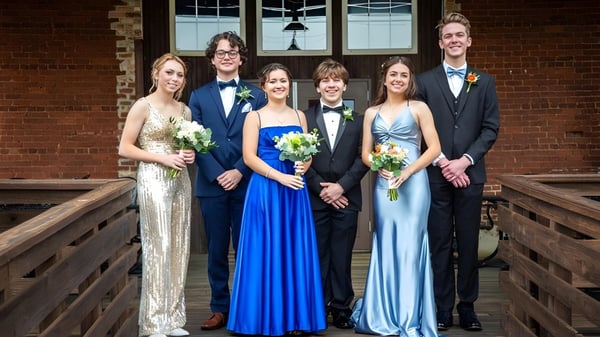 Un grupo de personas vestidas de forma formal se encuentra en un puente de madera frente a un edificio de ladrillo en el campus de la Springwood School.