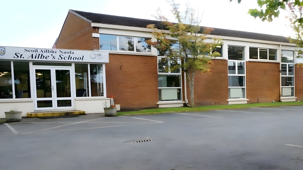 El edificio de ladrillo de dos pisos de St Ailbe's School con grandes ventanas se encuentra en un terreno pavimentado rodeado de árboles.