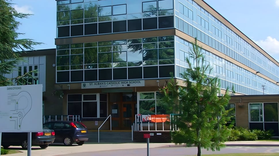 El moderno edificio de vidrio y acero de St. Alban's Catholic High School se encuentra bajo un cielo azul con árboles y un coche en primer plano.
