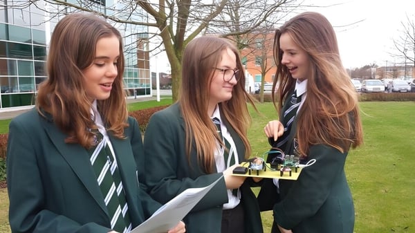 Tres estudiantes en uniformes escolares están en el terreno de St. Alban's Catholic High School con un edificio y árboles de fondo.