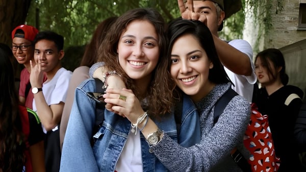 Dos estudiantes se abrazan sonriendo en un grupo en el área verde exterior del St. Andrew’s College en Cambridge.