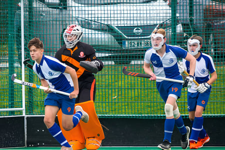 Un grupo de jóvenes jugadores de hockey con camiseta y un portero frente a una cerca de alambre verde en el terreno del St. Andrew's College.