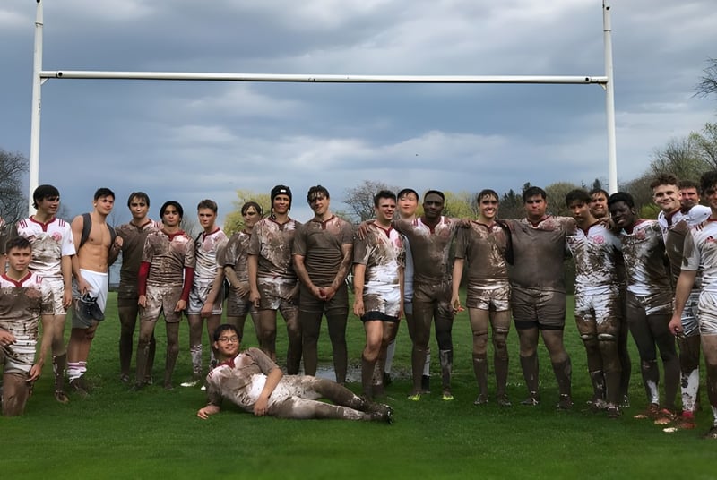 Un grupo de estudiantes cubiertos de barro está frente a una portería de rugby en el campo de deportes del St. Andrew’s College.