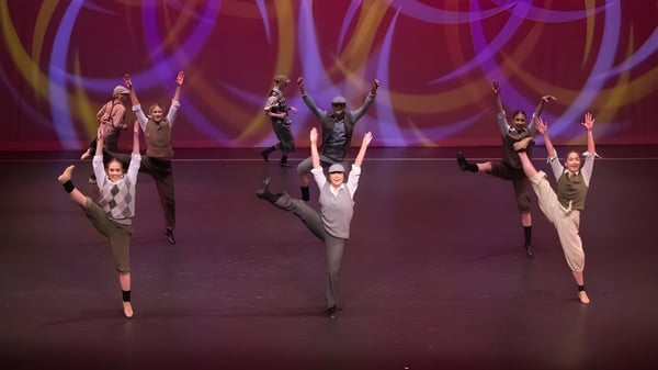 Un grupo de estudiantes del St. Andrew's College baila en el escenario frente a luces vibrantes en color púrpura y naranja.