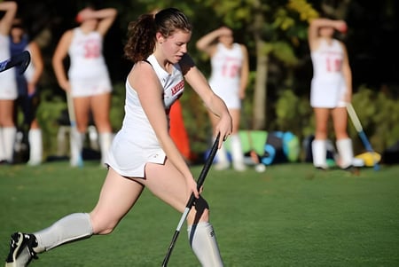 Una estudiante de la St. Andrew's School corre con ropa deportiva sobre un campo verde con otras jugadoras al fondo.