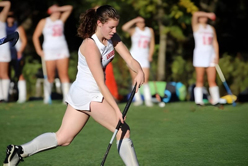 Una estudiante de la St. Andrew's School corre con ropa deportiva sobre un campo verde con otras jugadoras al fondo.