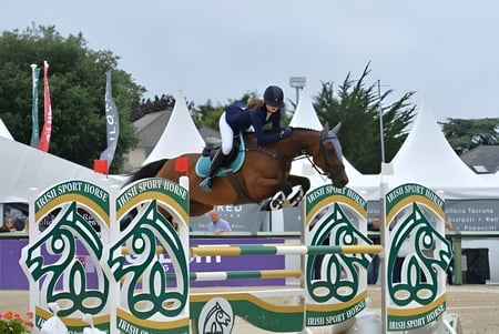Un estudiante de St. Anne's Secondary School salta con un caballo sobre un obstáculo colorido en un evento ecuestre al aire libre.
