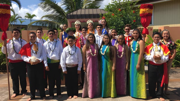 Un grupo de estudiantes de la St. Anthony School lleva ropa tradicional frente a un paisaje tropical con palmeras.