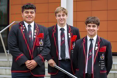 Tres estudiantes en uniforme escolar formal están frente a un moderno edificio naranja en el campus de St. Bede's College.