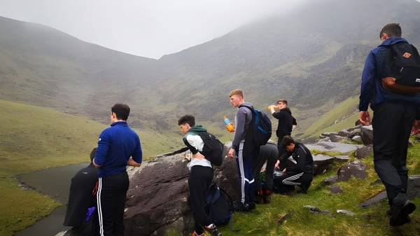 Estudiantes del St. Brendan's College están de pie en ropa deportiva sobre una roca con vista a un paisaje montañoso nublado.