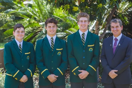 Cuatro estudiantes del St. Brendan's College están de pie en trajes verdes y morados en un área exterior tropical con palmeras.