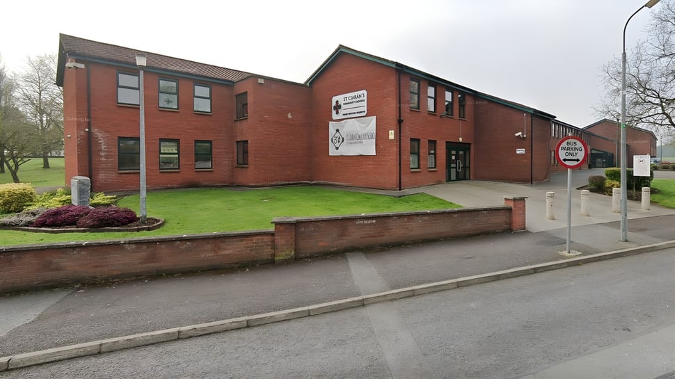 El edificio de ladrillo de dos pisos de la St. Ciarán’s Community School con varias ventanas se encuentra en una zona de césped frente a árboles y un cielo nublado.