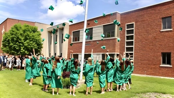 Un grupo de graduados de la St. Edmond Catholic School está de pie en togas verdes frente a un edificio de ladrillo con banderas y decoraciones.
