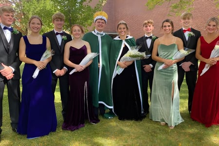 Un grupo de estudiantes vestidos formalmente está de pie en el verde frente a un edificio de ladrillo en el terreno de la St. Edmond Catholic School.