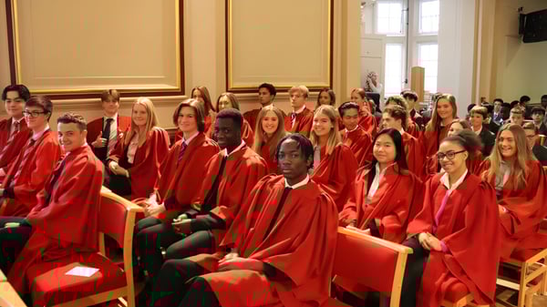 Estudiantes de la St. Edmunds School llevan togas rojas durante una ceremonia en una sala decorada con grandes ventanas.