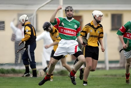 Un grupo de estudiantes juega hurling en el campo de deportes de St. Fergal’s College con edificios de fondo.
