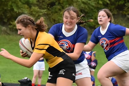 Tres alumnas del St. Finian’s College Mullingar practican deportes en un campo cubierto de hierba con árboles al fondo.