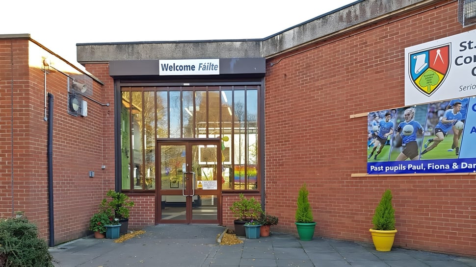 El área de entrada de St. Finian’s Community College con un cartel de Welcome Fitness y plantas frente al edificio de ladrillo.