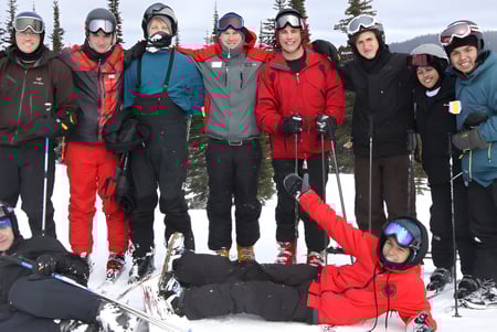 Un grupo de estudiantes de St. George’s School está de pie con colorida ropa de invierno en una ladera nevada con un bosque al fondo.