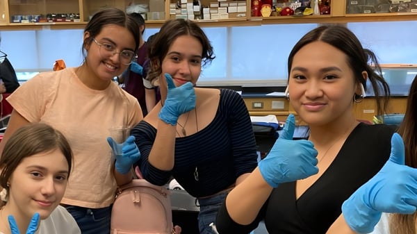 Cuatro alumnas de St. James Collegiate llevan guantes y posan juntas en un laboratorio con estantes y equipo.