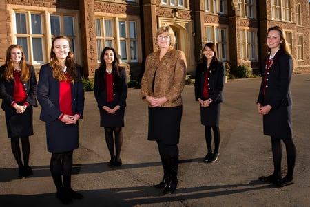 Cinco mujeres vestidas formalmente están juntas frente a un edificio histórico de ladrillo de la St. James’ School.
