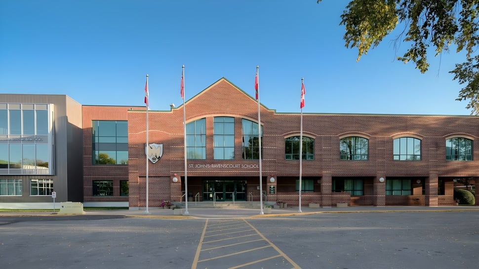 El gran edificio de ladrillo con ventanas de arco en el campus de la St. John’s-Ravenscourt School está rodeado de un área pavimentada y árboles.