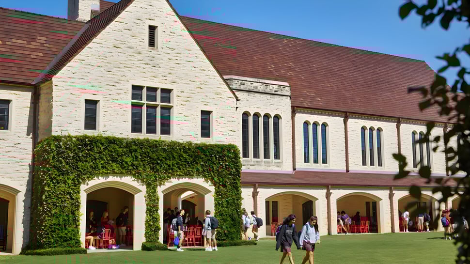 El gran edificio de piedra de la St. John's School está rodeado de un exuberante verde y en primer plano hay personas sentadas en bancos.