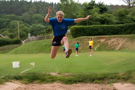 Una persona con camisa azul salta alto en un campo frente a un bosque en el terreno de la St. John’s School Sidmouth.