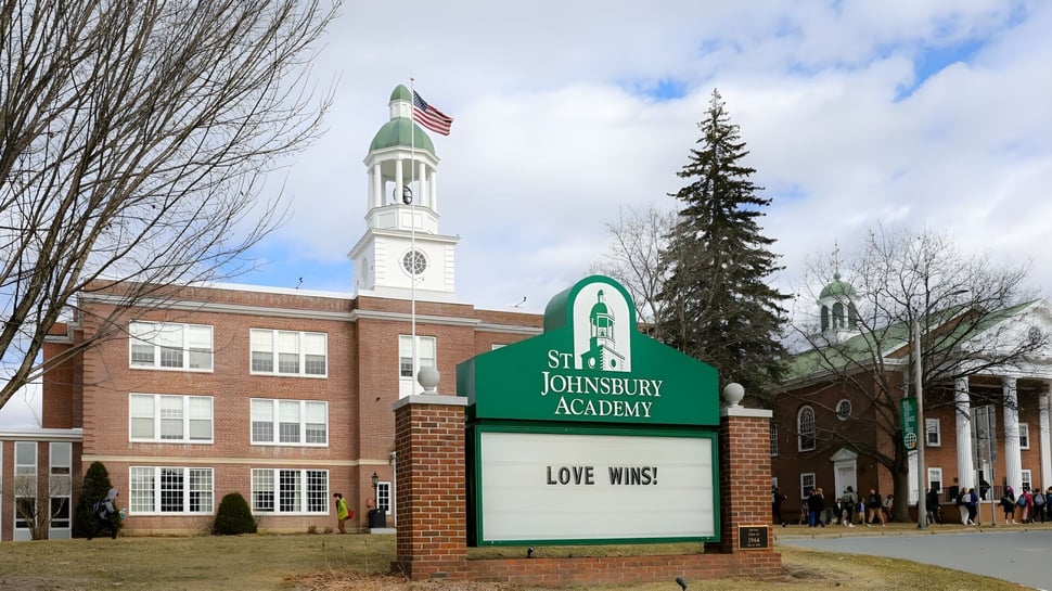 El edificio de ladrillo con torre del reloj y bandera americana en el campus de la St. Johnsbury Academy rodeado de árboles y césped.
