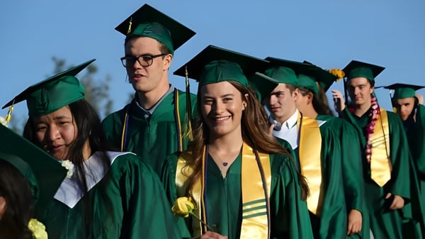 Un grupo de graduadas y graduados de la St. Joseph Academy está frente a un cielo azul radiante con togas verdes y amarillas.