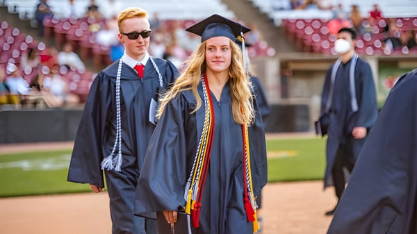 Una graduada en toga y birrete está frente al campus de la St. Joseph High School con otros graduados al fondo.