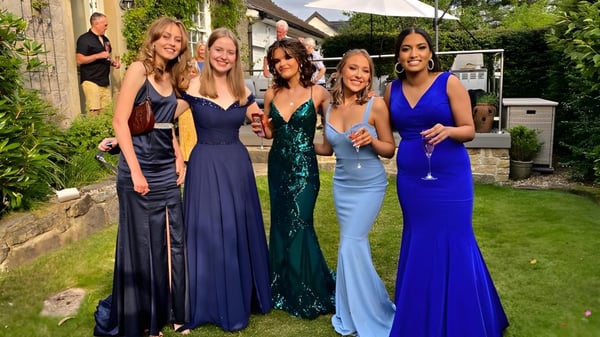 Cinco estudiantes del St. Leo’s College están en elegantes vestidos de noche en el jardín frente a un muro de piedra.